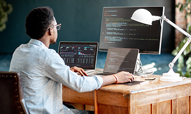 back of person coding at desk with laptop, monitor and tablet
