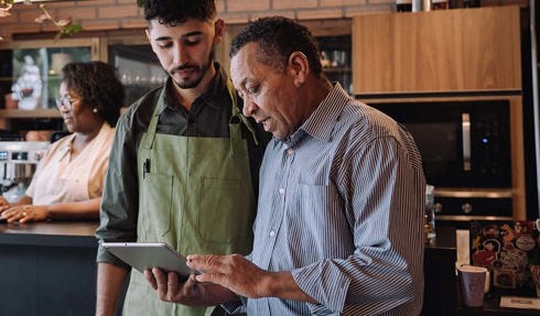 Two people reviewing information on a tablet in a café or small business setting, with another person working on a laptop in the background.